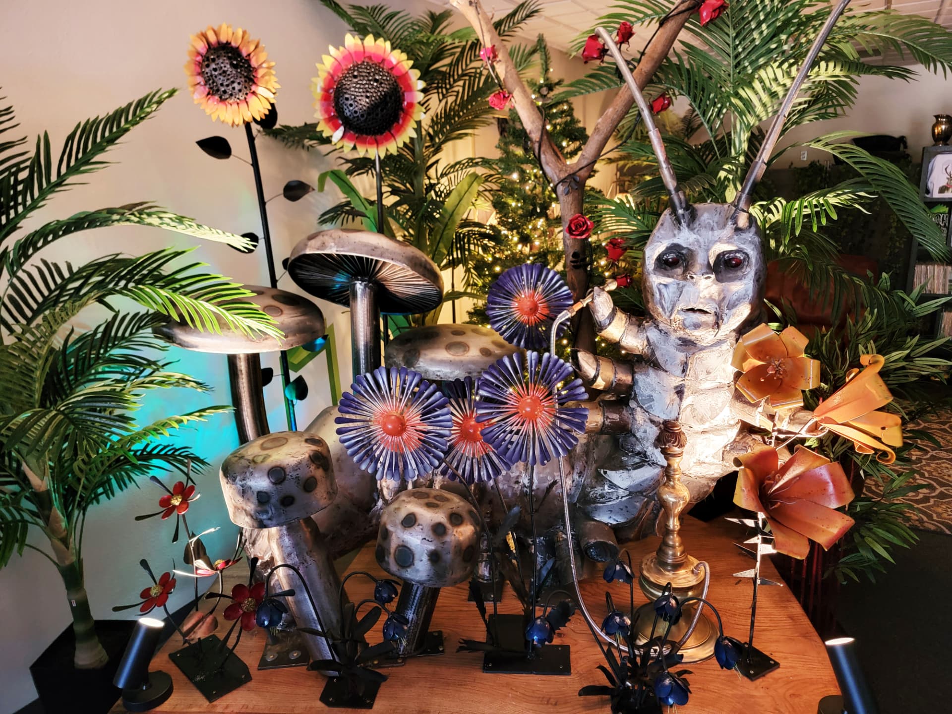 Gallery installation of salvaged-metal mushrooms, sunflowers, flowers and a large seated caterpillar figure arranged among plants and spotlights