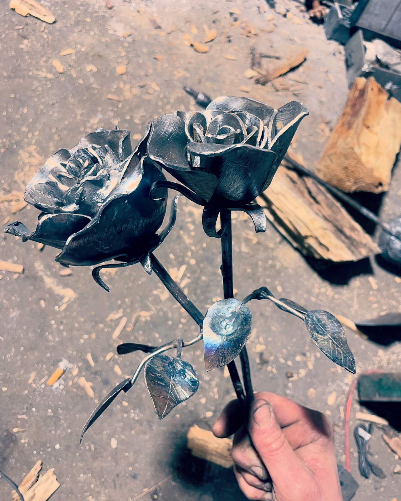Hand-forged bare steel double rose sculpture with veined leaves, held up in the workshop