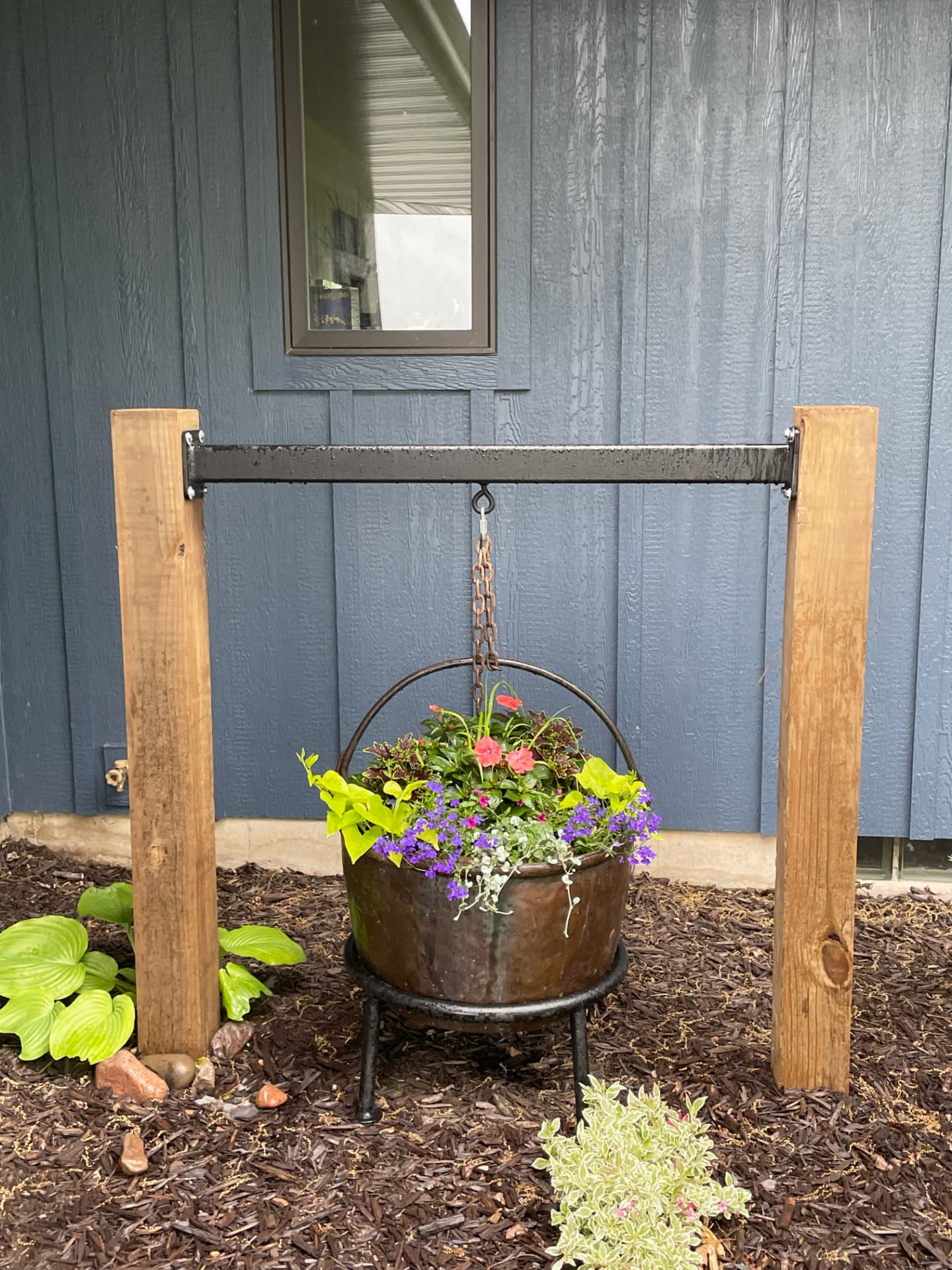 A hanging planter with pink and purple flowers suspended by chain from a steel bar mounted between two cedar posts, set in a mulched garden bed against a blue house wall